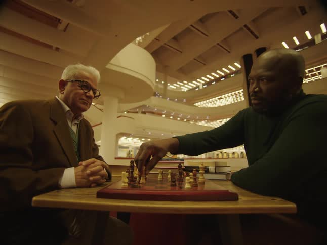 Two men are sitting at a table inside an elaborate building playing chess