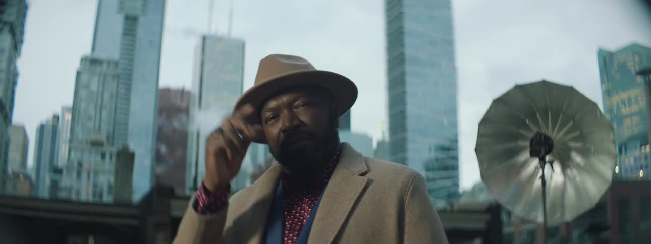 Male model wearing a suit and a hat stands with city as a backdrop as he moves through poses