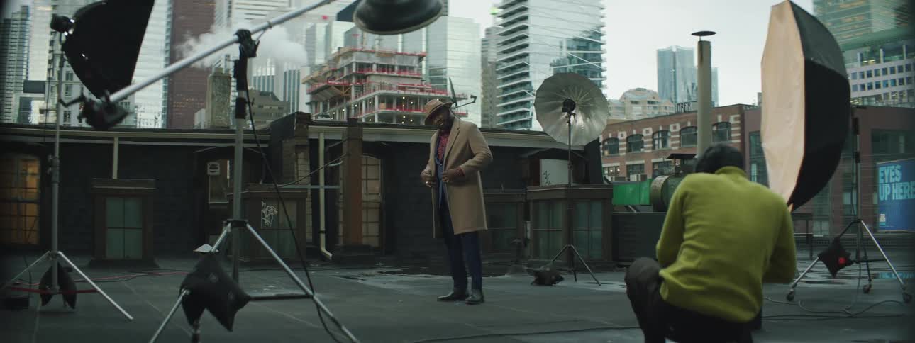 A black man is on a stage modeling a suit and there are tall city buildings behind the stage