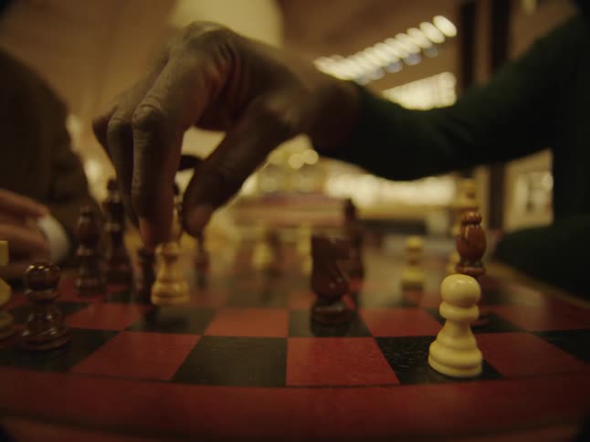 A man hesitantly moves a chess piece on a chess board as his opponent shakes his head and smiles