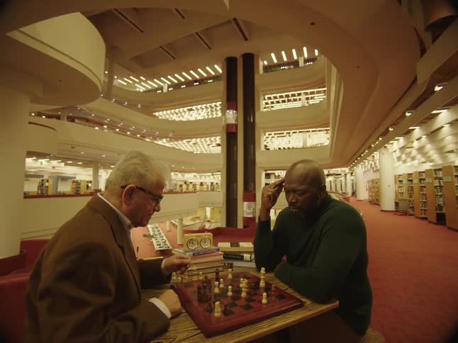 Two men play chess at table in large multi story library as elevators move up and down in background