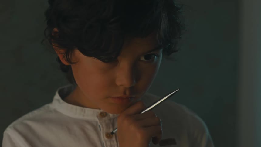 A child is standing in a room holding a pen and staring off