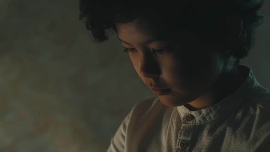 Close up of a boy wearing a white shirt writing inside a dimly lit room at home
