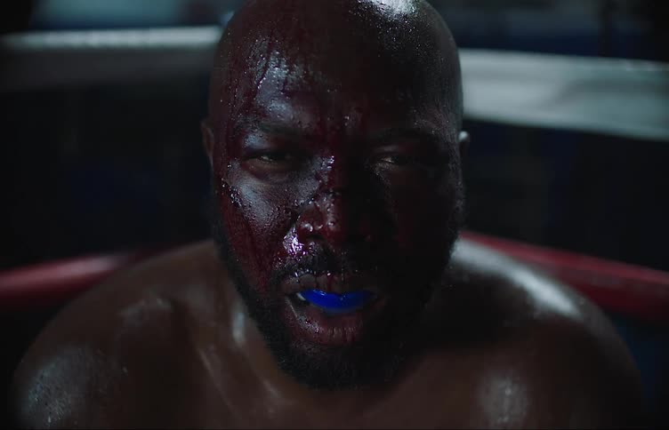 Bald boxer with blue gumshield sweat and bloody face sits in the corner of ring