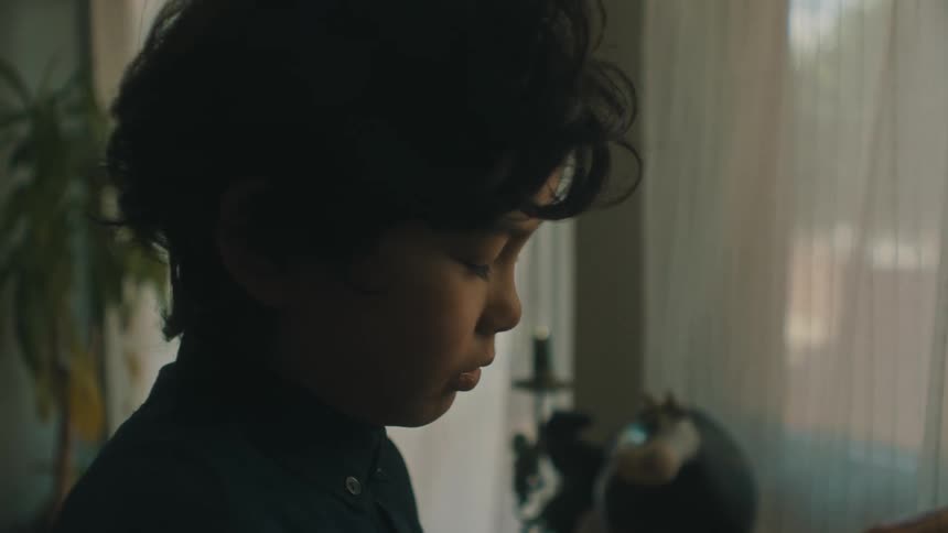 A boy stands near windows at home looking down and sighing