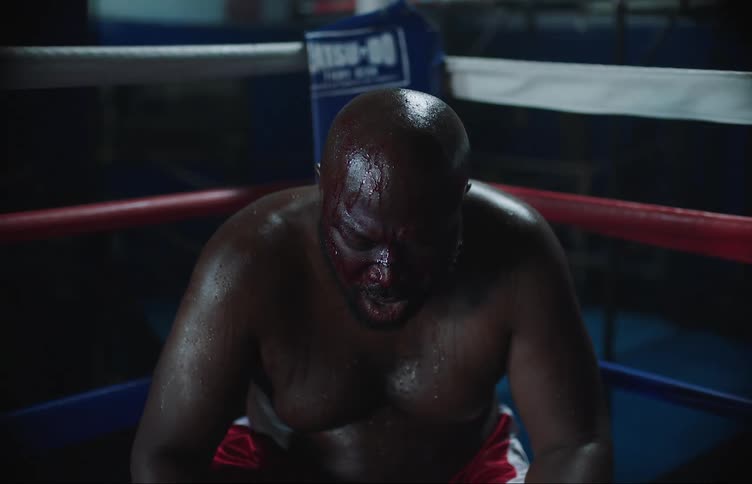 A heavyweight boxer sitting in his corner panting covered in sweat with blood on his face