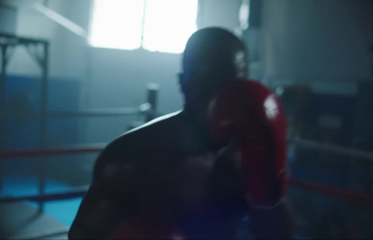 Strong muscular man inside a ring on a dimly lit gym boxing at the camera