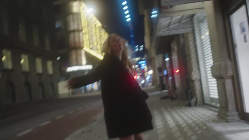 A woman with long blond hair wearing a dress and a fur coat is dancing on a city street at night