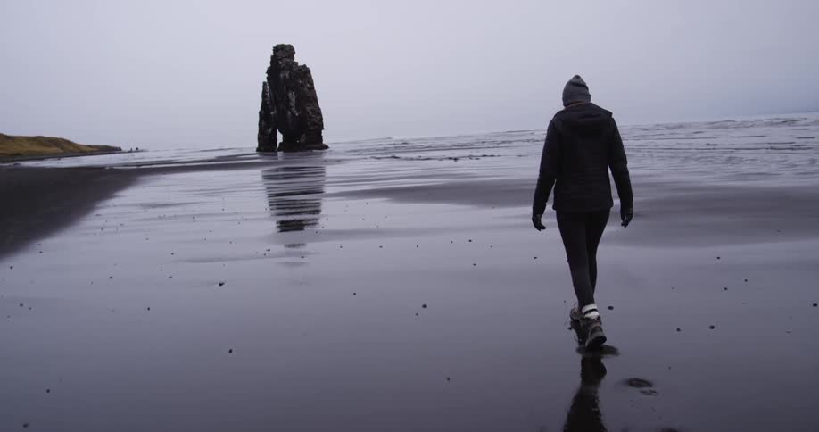 A woman is walking on the shore towards some rocks