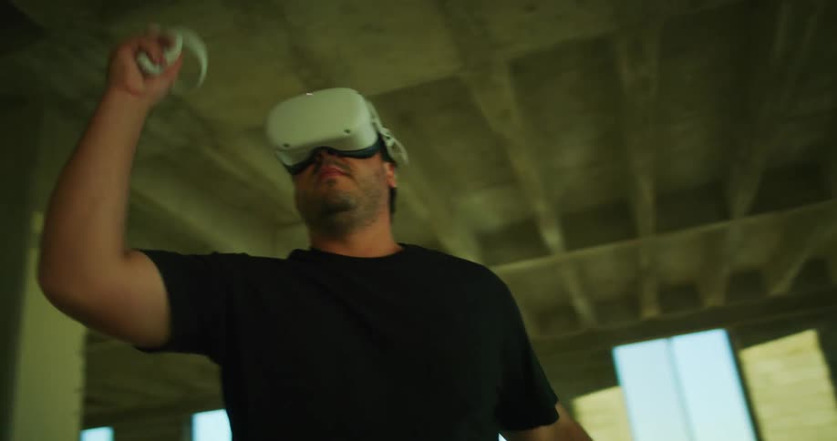 A man wearing a virtual reality headset has a gaming controller in each hand as he swings his arms playing a game