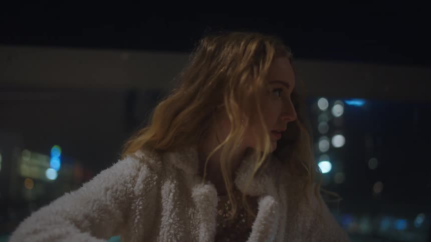 Woman with brown wavy hair white feather jacket uses smartphone and looks around in city street at night