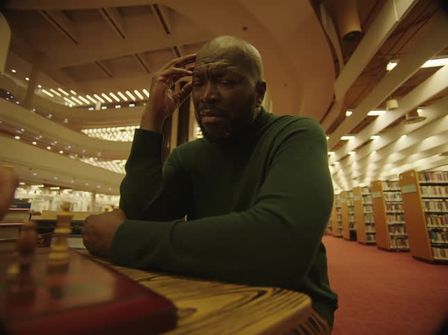 A man is sitting with his fingers against his head as he watches another person move a chess piece