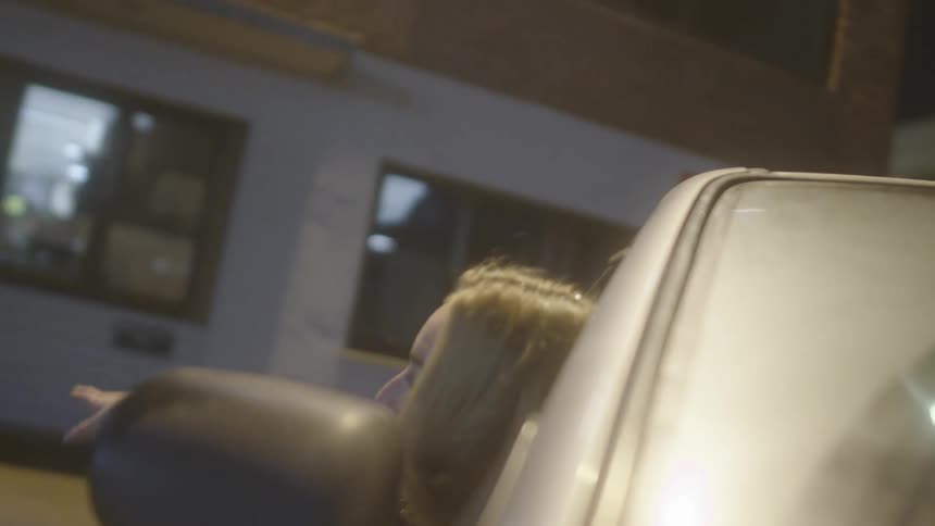 A woman smiles while sticking her head and arm out of the passenger side window of a moving car in a city at night