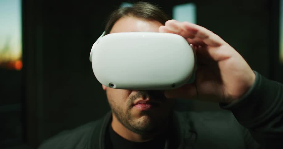 Man looks at the camera while wearing virtual reality goggles inside a room at sunset