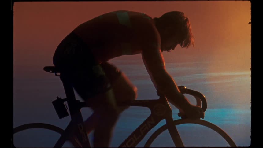 A man works out on an indoor bicycle in front of a screen displaying a sunset