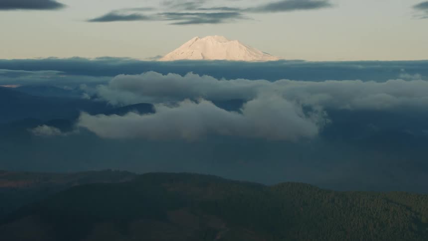 An aerial view of the landscape with clouds above a forest with a snow covered mountain in the distance