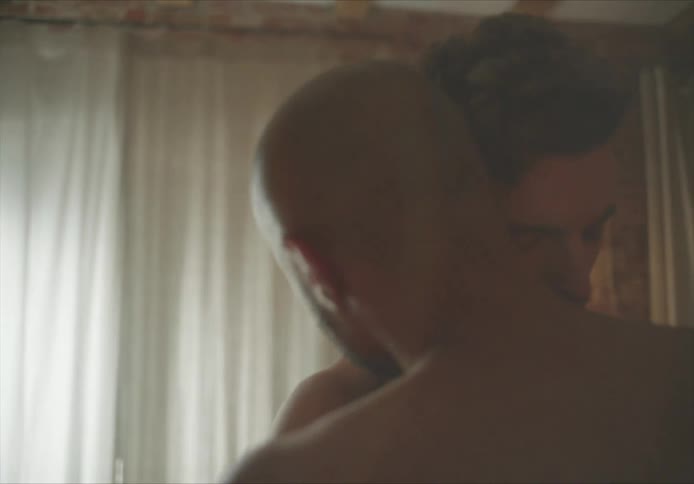 Two men slow dance bare chested in home near windows