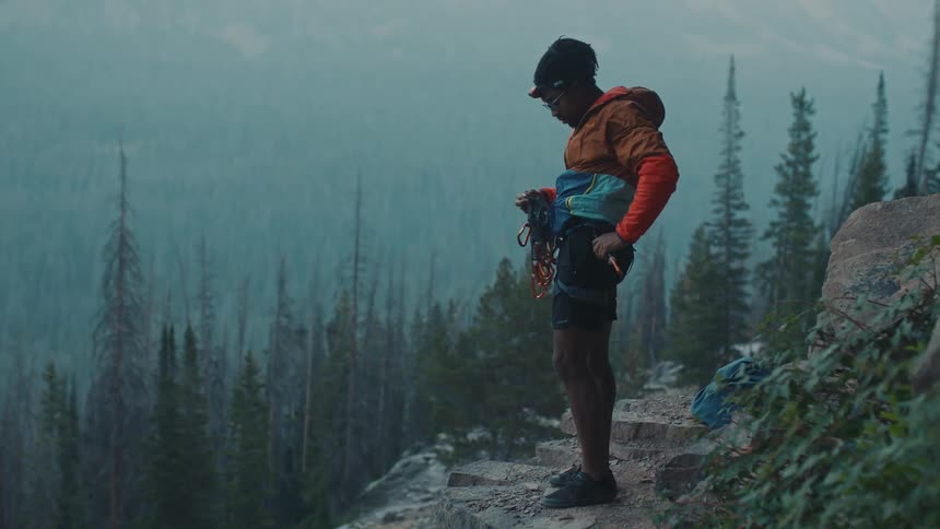 Man stands on mountain side overlooking fog obscured forest working with his climbing gear