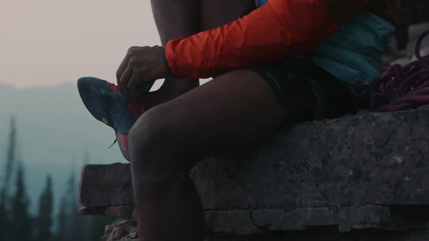 A man is sitting on a mountain ledge and he puts a shoe on his bare foot