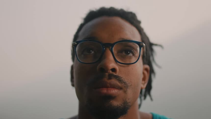 Young man wearing blue glasses and dreadlocks looks up standing outdoors