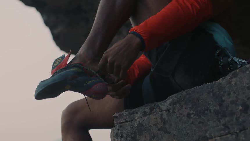 A rock climber is sitting on the cliff putting on his shoes