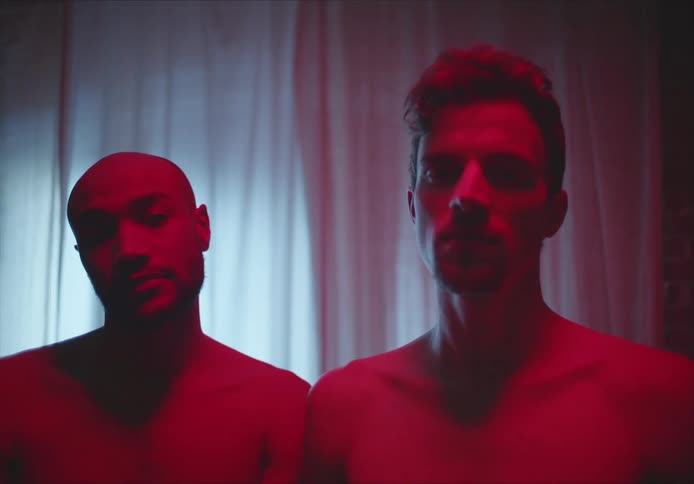 Two men stand together in front of a window in a home without shirts on
