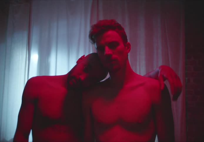 Two men stand under red lighting leaning against each other then hugging