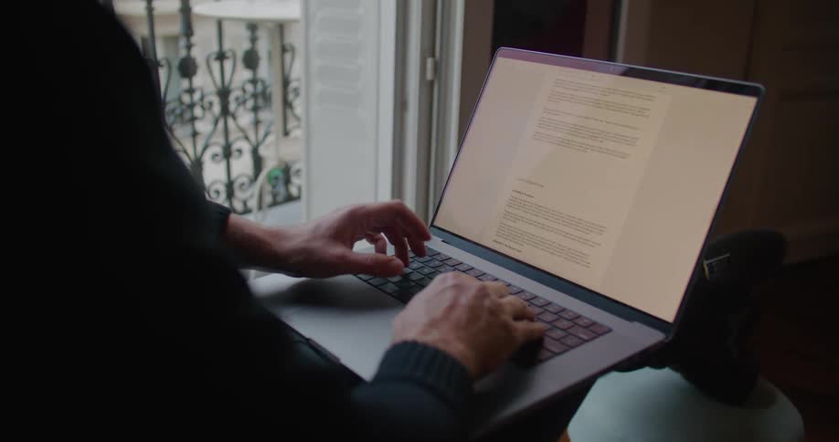 Man sits near sliding glass doors with his feet up and types on laptop in his lap