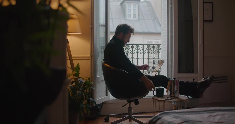 A man sits at home and drinks coffee as he types on his laptop