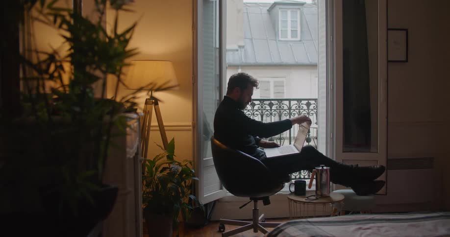 A man carries his laptop to a chair in his home and sits down and opens it on his lap and starts typing