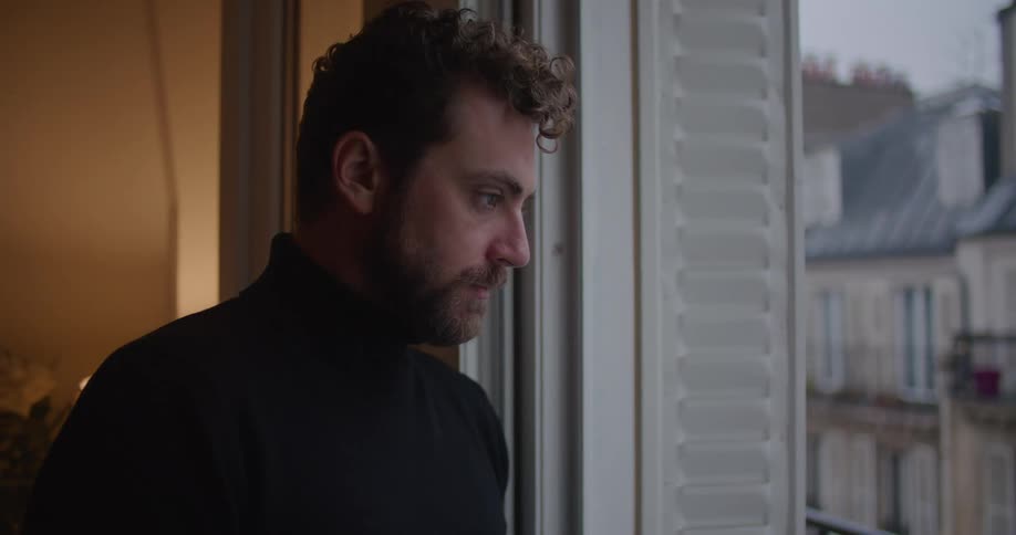 Man stands near door to balcony looking downcast before turning to go inside