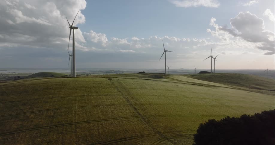 Wind turbines on top of grass covered hills turn slowly against the cloudy sky backed by misty red tinted horizon
