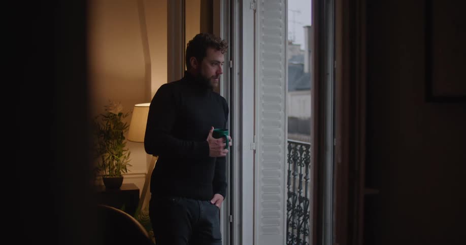Young man wearing a black sweater holding a mug looking out the apartment balcony