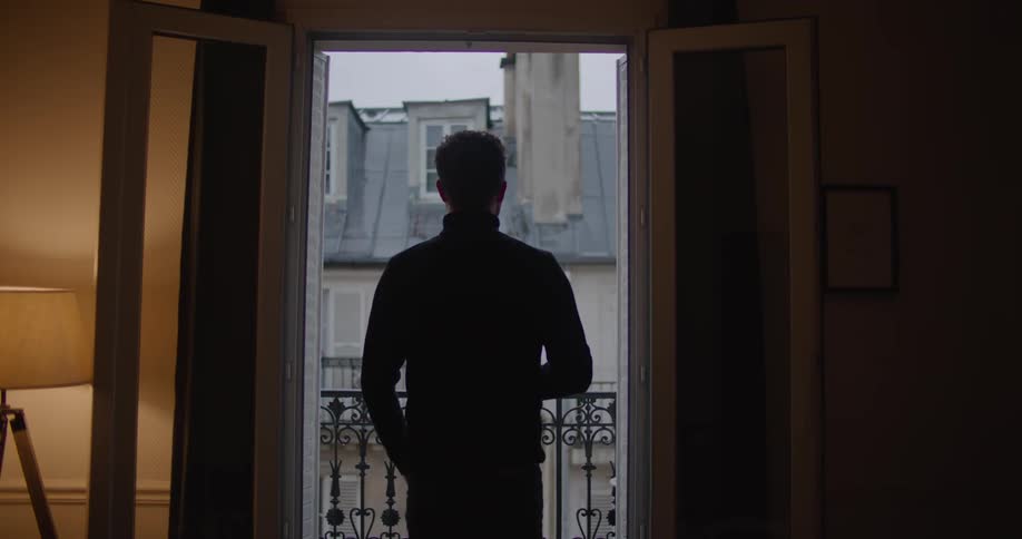 A man stands and looks out his balcony window and then walks away