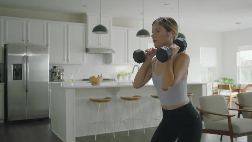 A woman holds dumbbells and does squats in her kitchen