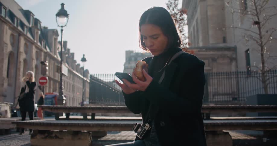 A young woman sits on a bench in a city and looks at her cell phone as she eats a pastry as a bird walks by her feet