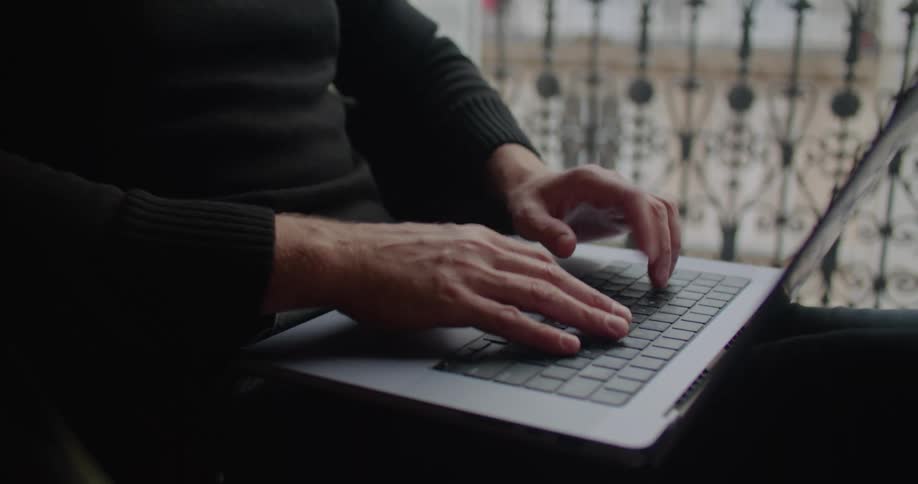 Man with beard and black turtleneck sweater uses laptop in apartment building next to balcony