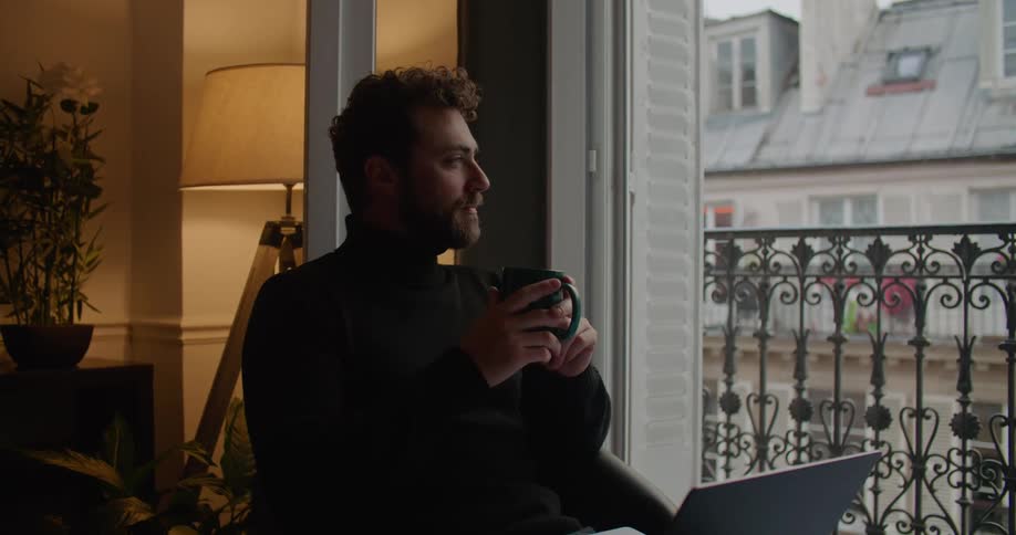 A man is sitting down inside with a cup in his hands and looking out the window