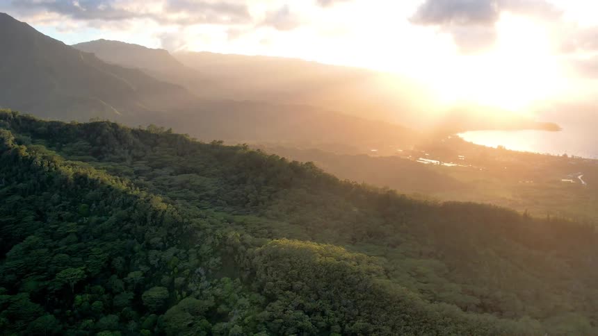 An aerial view of the sun setting over a hilly forest on the shore