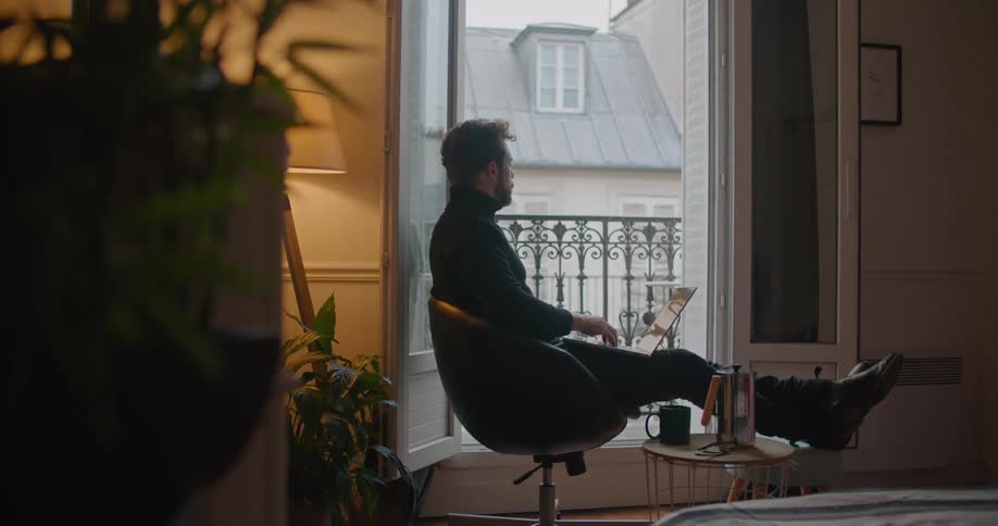 Man sits with feet up typing on laptop in his lap with balcony shown through open doors in the background