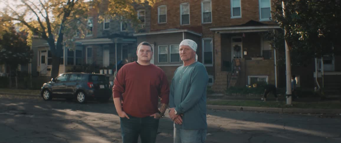 Two men pose in middle of road as kids in background play near houses