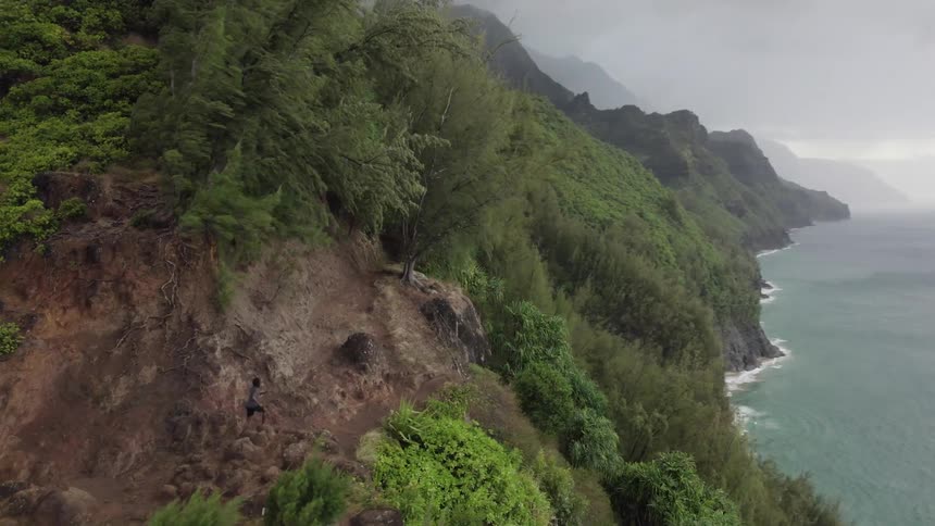 Aerial view of man with gray t shirt black shorts and shoes run on the edge of cliff overlooking shoreline on cloudy day
