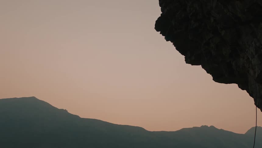 A person rock climbing down the edge of a mountain looks down as they descend