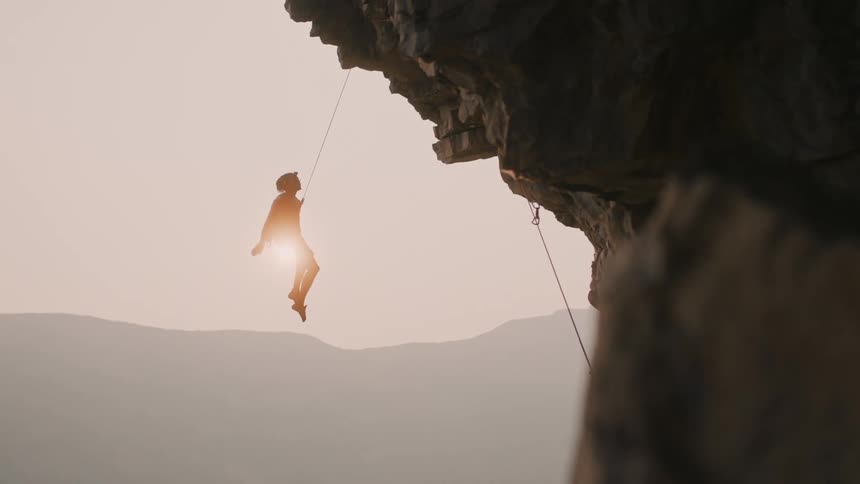 Young man climbing a mountain lets himself fall down while balancing on a rope at sunset