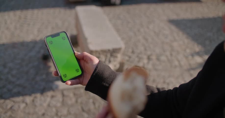 Woman eating a croissant in the park holding a smartphone with green replaceable screen