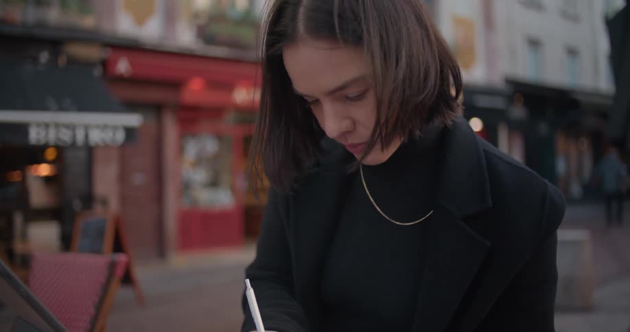 A woman sits at an outdoor cafe table writing in a notepad