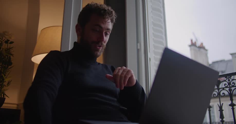 Man with beard turtleneck sweater takes a green cup to drink while using laptop in apartment building
