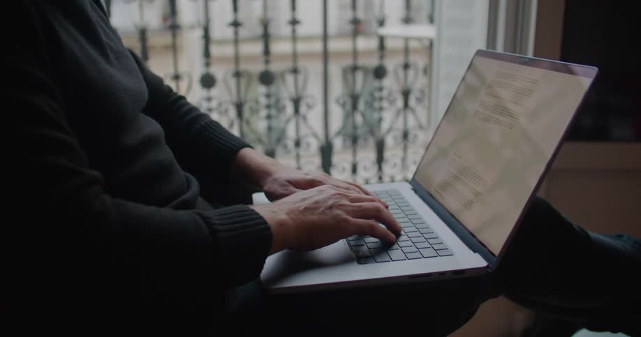 Man with beard and black sweater sitting in armchair drinks from green cup working on laptop in apartment building