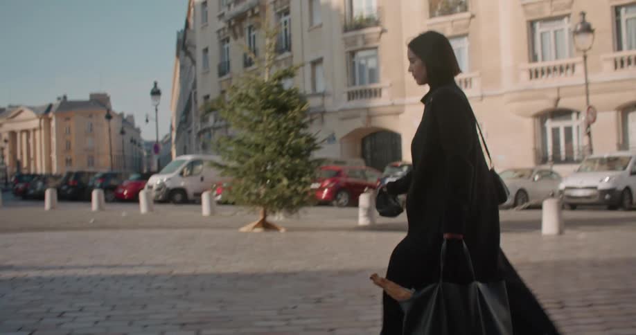 Woman with short straight hair and black topcoat walks down the street in the city
