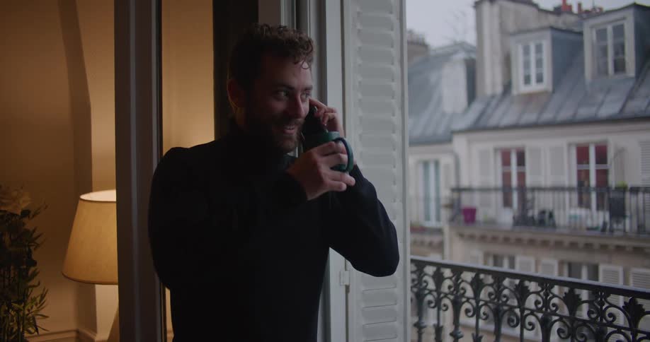 Man with beard black turtleneck sweater drinks from green cup and talks on smartphone looking out balcony door in apartment building
